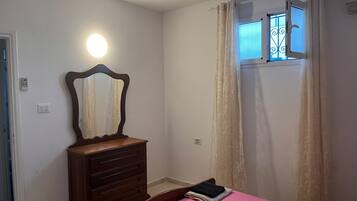 A bedroom with a bed, a dresser, a mirror, a window with curtains, and a wall-mounted light.