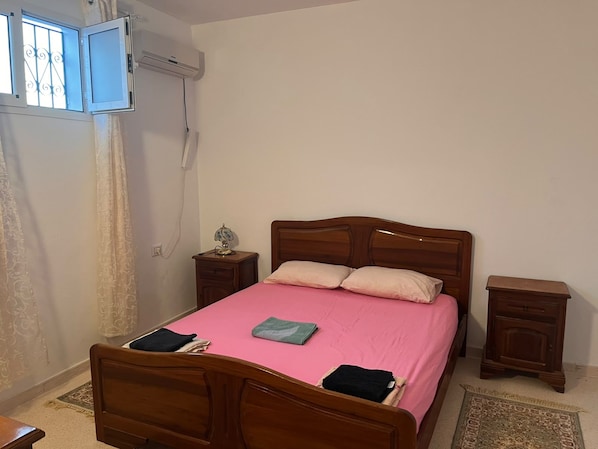 A bedroom with a wooden bed, a bedside table with a lamp, a small wooden nightstand, and a window with bars.