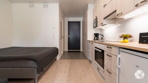 Quarto de hotel compacto com cama, cozinha compacta e porta que dá para um corredor escuro.