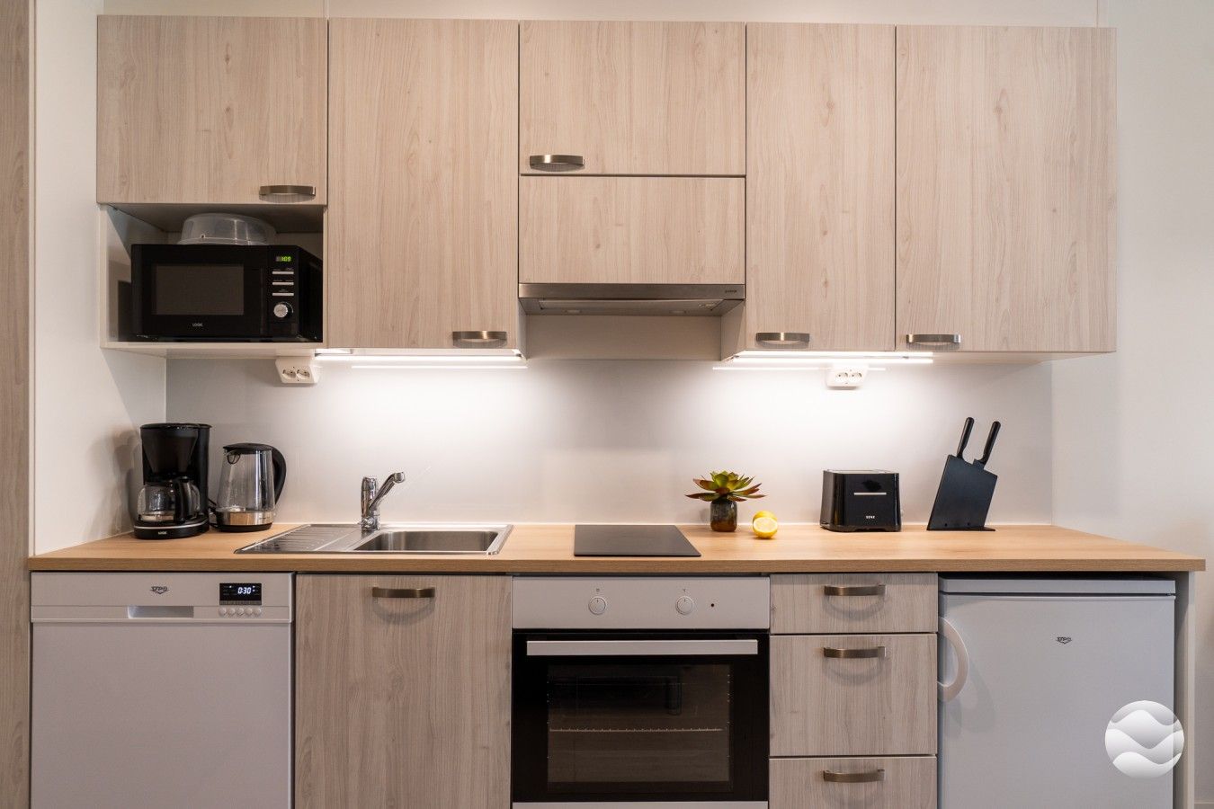 Una cocina moderna con armarios de madera, microondas, cafetera, pileta y un pequeño macetero, además de un bloque de cuchillos.