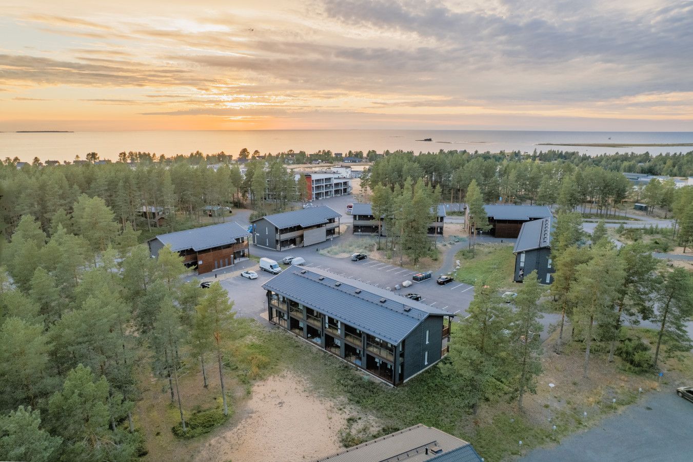 Moderni rakennuskompleksi, jossa on useita rakennuksia, ja se sijaitsee tiheän metsän ympäröimänä, taustalla näkyy vesistö.