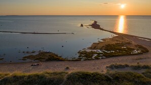 Paisagem costeira com o pôr do sol refletindo na água, uma praia arenosa e uma pequena península com vegetação.