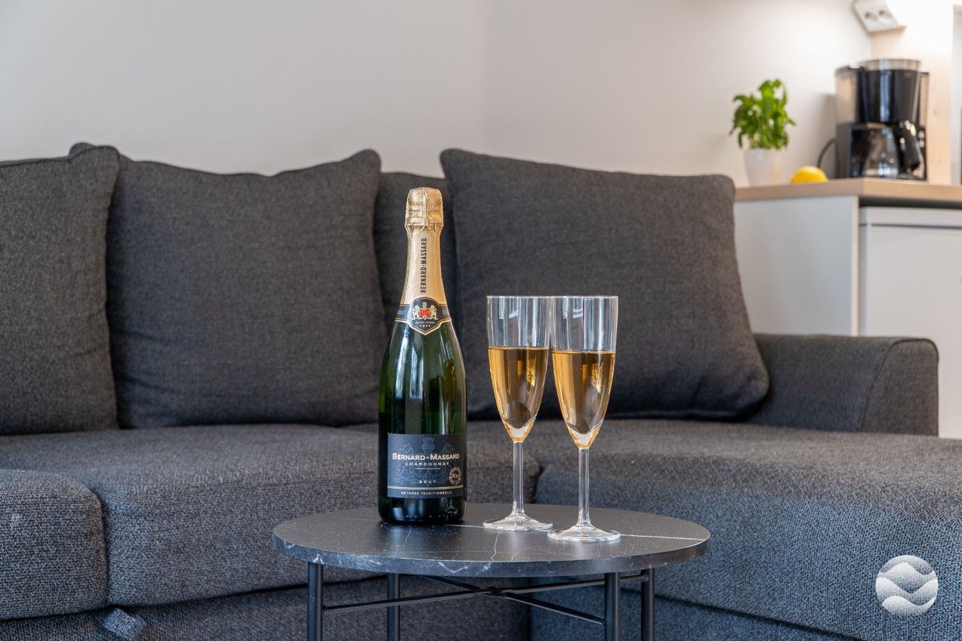 Bernard-Massard Chardonnay -pullo ja kaksi viinilasia pienellä pyöreällä pöydällä harmaan sohvan edessä.