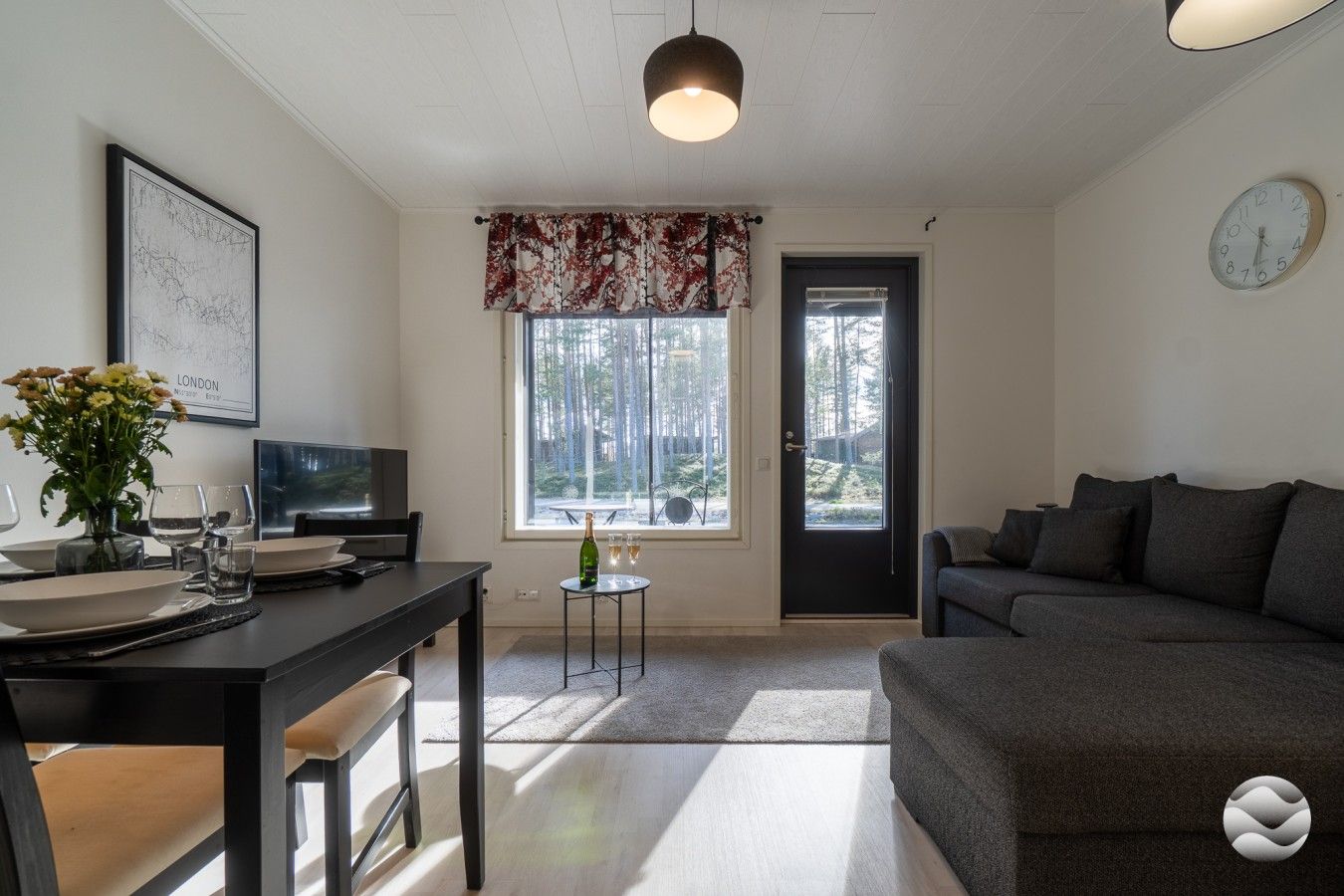 Uma sala de estar moderna com um sofá escuro, uma área de jantar com mesa posta para dois e uma janela grande com vista para árvores.