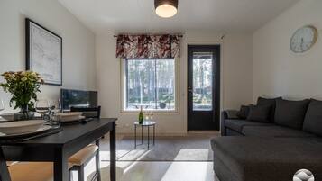 Uma sala de estar moderna com um sofá escuro, uma área de jantar com mesa posta para dois e uma janela grande com vista para árvores.