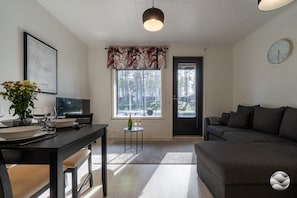 A modern living room with a dark sofa, a dining area with a table set for two, and a large window with a view of trees.
