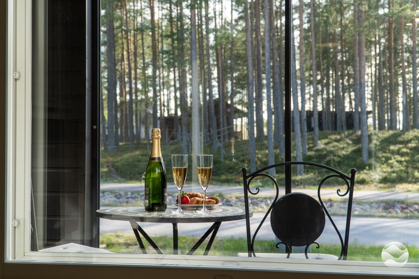 Pöydällä on pullo ja laseja, sen vieressä tuoli ja ikkunasta avautuu näkymä metsään.