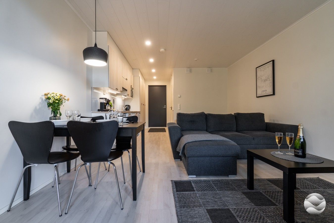Uma sala moderna com sofá, área de jantar com cadeiras, cozinha ao fundo e uma pequena mesa com copos e garrafa.