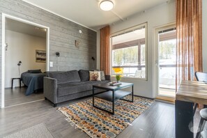 A modern living room with a grey sofa, a coffee table, and a patterned rug. There is a window with curtains, a door leading to another room, and a small table with a plant. - Polar Charm G Holiday in Lapland (Ylläs)