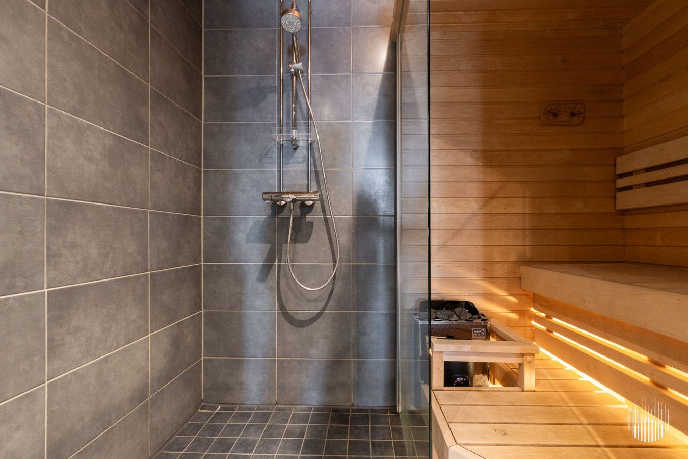 Una sauna moderna con una panchina in legno, cabina doccia in vetro e stufa a legna visibile.
