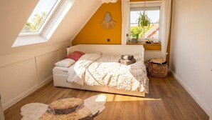 Un dormitorio con techo inclinado, una cama con edredón blanco, un ventanal con cortinas y una maceta sobre el alféizar.