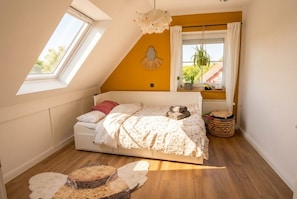 A bedroom with a sloped ceiling, a bed with a white duvet, a small window with curtains, and a potted plant on the windowsill. - Vrbo Property (Ahlerstedt)