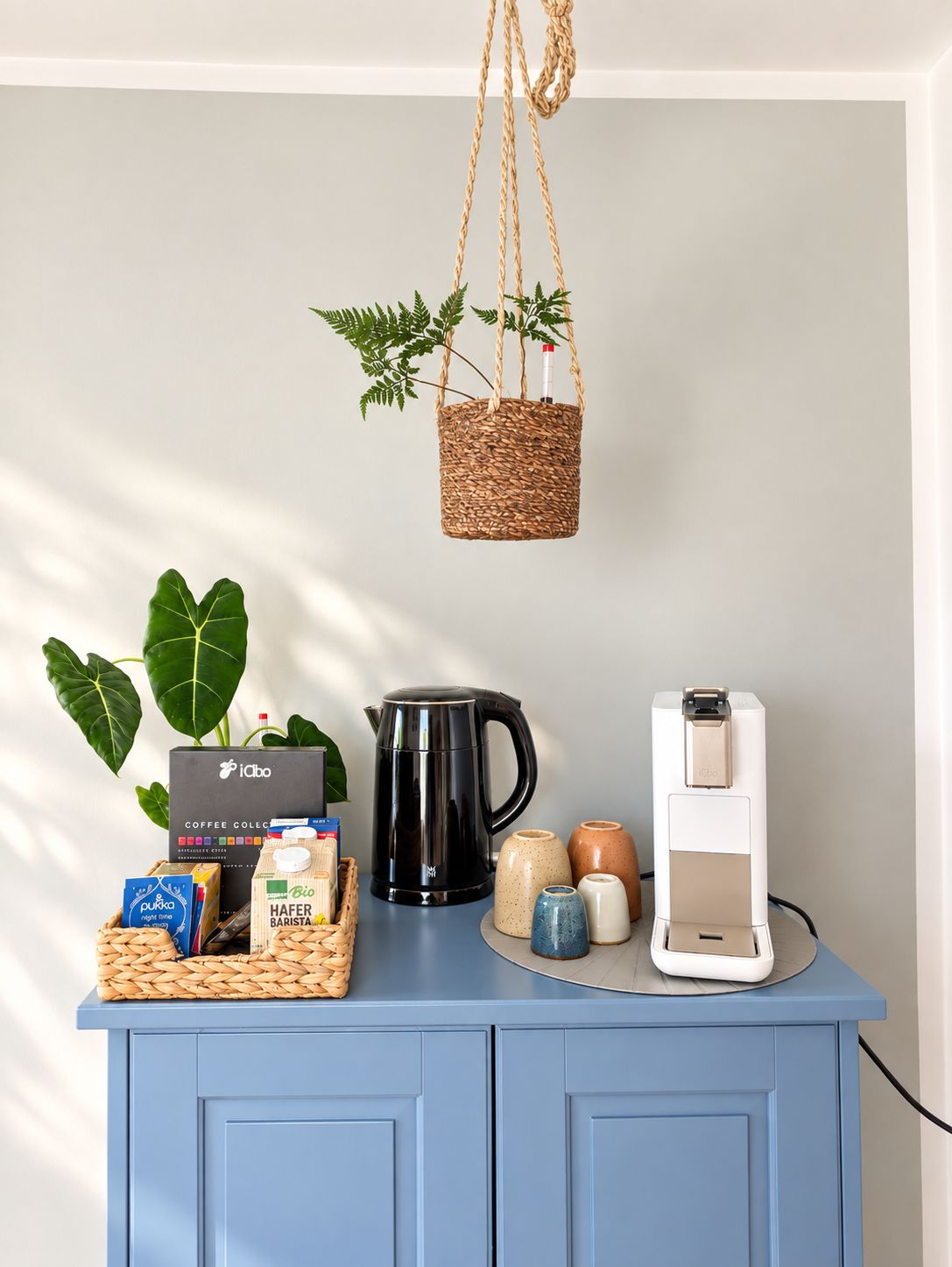 A kitchen counter with a coffee maker, a coffee pod dispenser, a basket of coffee pods, a coffee grinder, and a hanging plant.