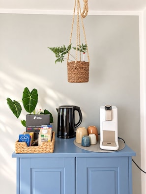 A kitchen counter with a coffee maker, a coffee pod dispenser, a basket of coffee pods, a coffee grinder, and a hanging plant.