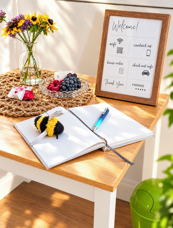 A wooden table with a framed welcome sign, a notebook, a pen, a bee plush toy, a vase with flowers, and a basket of berries. - Vrbo Property (Ahlerstedt)
