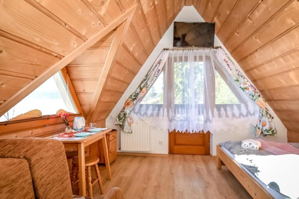 A cozy attic bedroom with a wooden sloped ceiling, a small table with chairs, a bed, and a window with lace curtains.