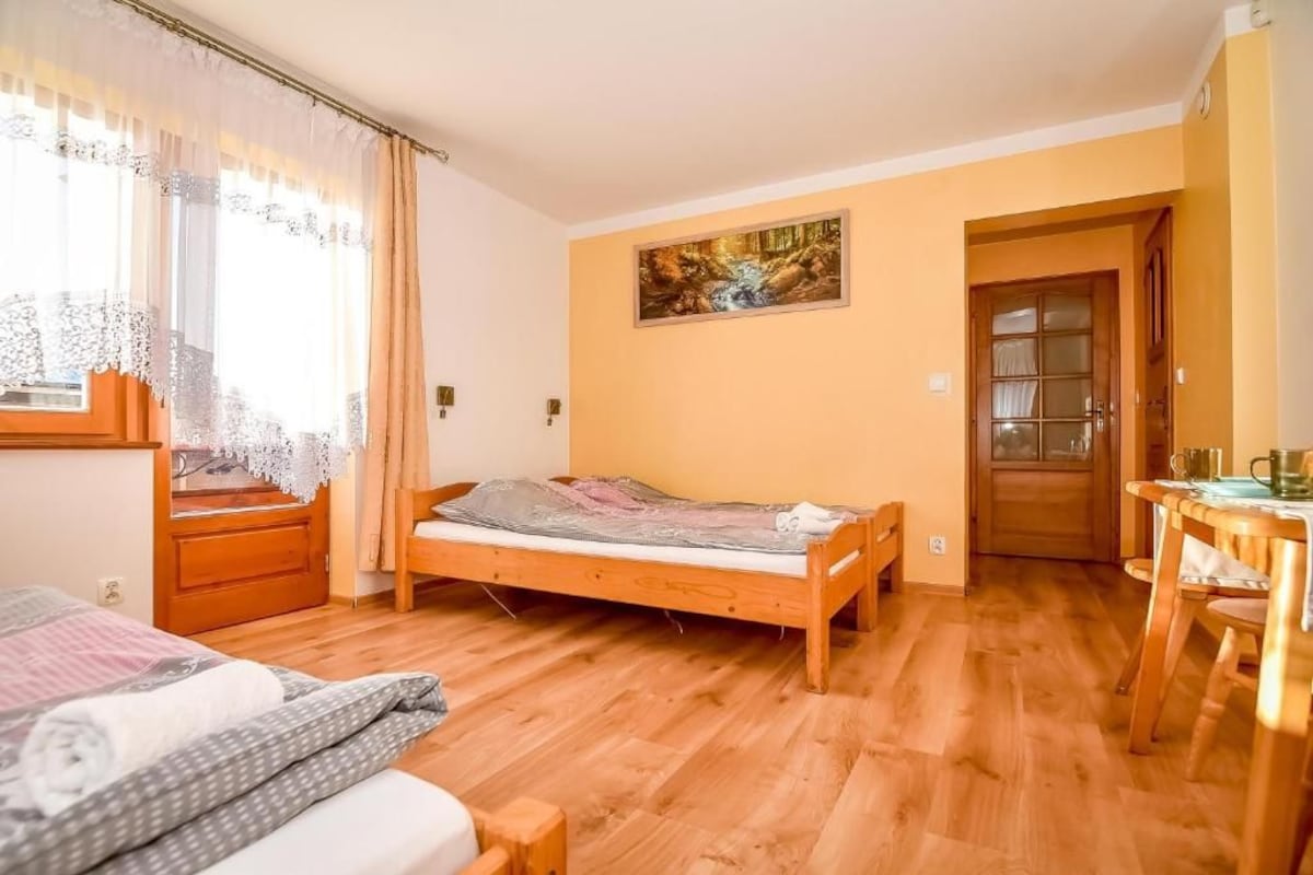 A bedroom with a wooden bed, a window with curtains, a painting on the wall, and a wooden door.