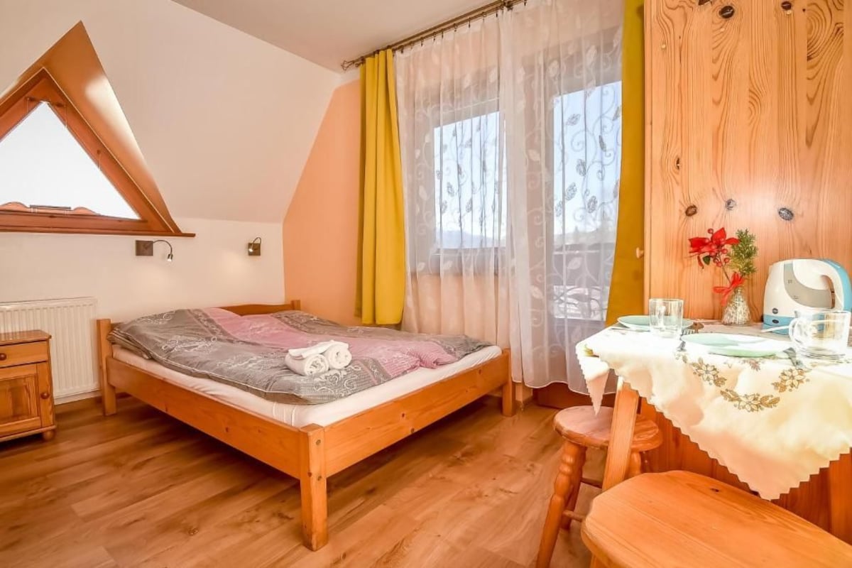 A bedroom with a sloped ceiling, a wooden bed, a table with a white tablecloth, and a window with sheer curtains.