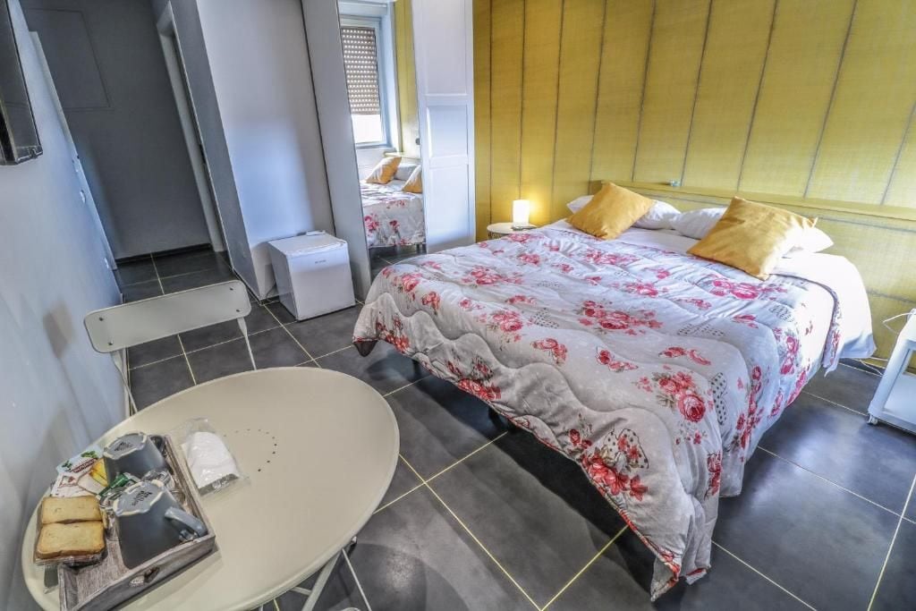 Une chambre d’hôtel avec un lit, une table de chevet et une chaise. On y trouve une petite table avec des aliments et une bouteille. La chambre a un sol carrelé et une fenêtre avec des stores.