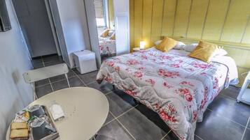 Une chambre d’hôtel avec un lit, une table de chevet et une chaise. On y trouve une petite table avec des aliments et une bouteille. La chambre a un sol carrelé et une fenêtre avec des stores.