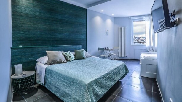 A modern bedroom with a large bed, a small round table, a chair, and a view of the city through a window. - IL GRATTACIELO B&B (Napoli)