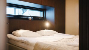 A bed with white bedding, a window above it, and a bedside table with a cup and a small container.