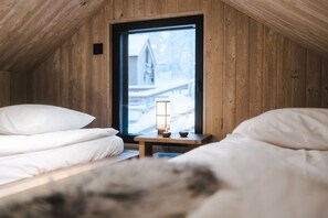 A cozy room with wooden walls and a large window. There are two beds with white bedding and a small table with a lamp.