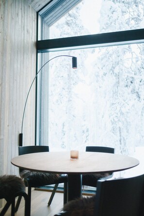 A round table with chairs, a candle, and a floor lamp in a room with wooden walls and a large window.