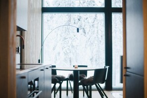Un espace repas moderne avec une table ronde et des chaises, un luminaire suspendu et une vue sur un paysage enneigé grâce à de grandes fenêtres.