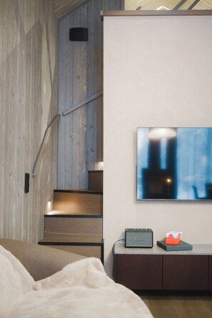 Une chambre à coucher avec un lit, un téléviseur fixé au mur, une petite enceinte et un escalier menant à un niveau supérieur.