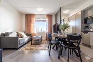 A modern living room with a sofa, dining table, and chairs, a kitchen area with appliances, and a large window with curtains. - Polar Charm C by Holiday in Lapland (Ylläs)