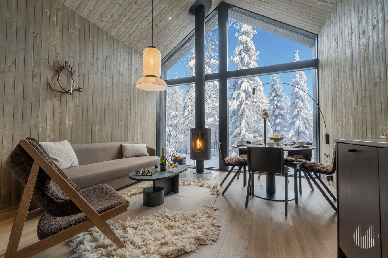 A cozy living room with a fireplace, a sofa, a dining table, and a large window with a view of snow-covered trees.