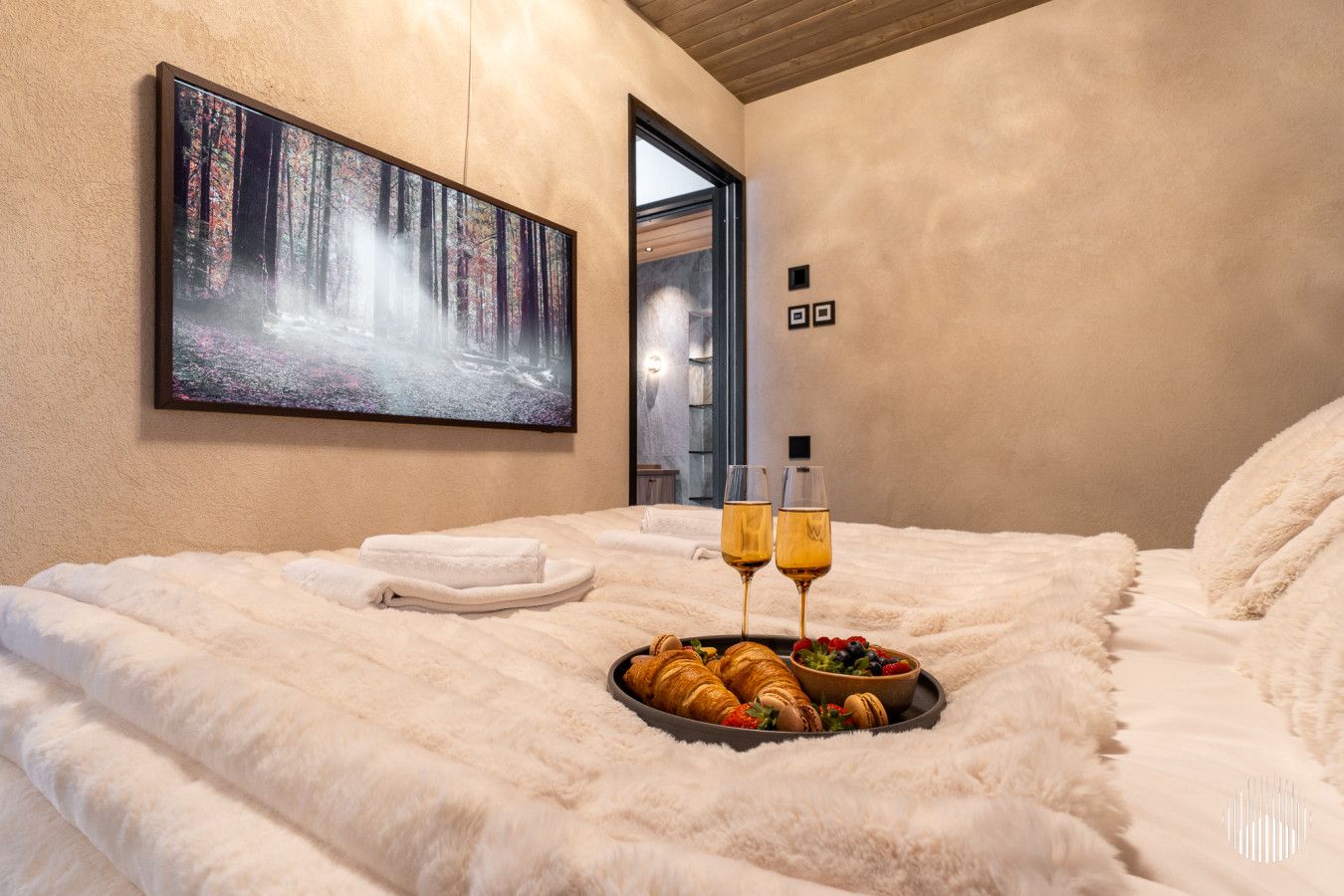 Une chambre avec un grand lit, un tableau représentant une forêt et un plateau avec des croissants et des fruits.