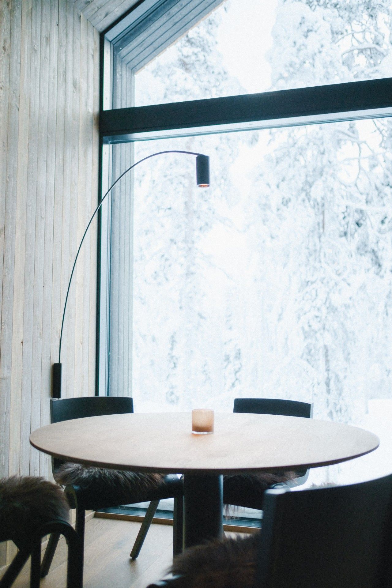Une table ronde entourée de chaises, une bougie et un lampadaire dans une pièce aux murs en bois et dotée d’une grande fenêtre.