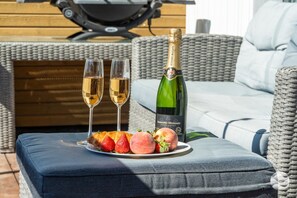 A wicker sofa with a cushion, a bottle of Bernard Massard Chardonay wine, two glasses of wine, and a plate with strawberries and peaches.