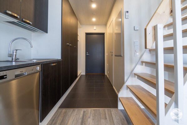 A modern kitchen with a dishwasher, a stainless steel sink, and a wooden staircase leading to a door.