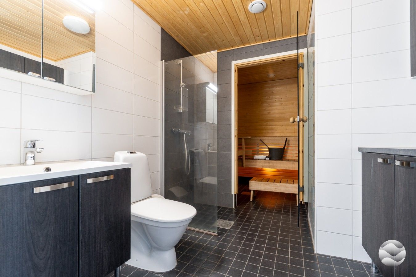 Moderni kylpyhuone, jossa on valkoinen wc-istuin, tumman puun väriset kaapit, lasinen suihkuseinä ja sauna, joka näkyy avoimesta ovesta.