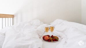 Cama com lençóis brancos, um prato de frutas e dois copos de uma bebida transparente.