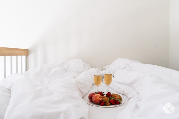 Un lit avec du linge de lit blanc, une assiette de fruits et deux verres contenant une boisson claire.