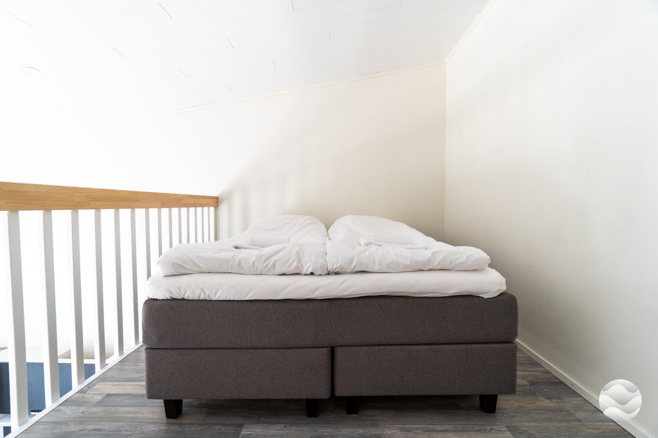 Een bed met een grijze matras en witte beddengoed in een kamer met witte muren en een houten plafond.