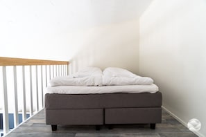 Un lit avec un matelas gris et du linge de lit blanc, dans une pièce aux murs blancs et au plafond en bois.