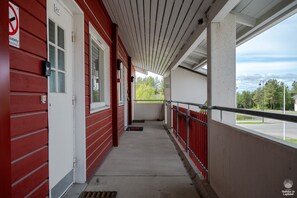 Ein überdachter Gang mit roter Außenfassade, einer weißen Tür und einer Straßen- und Baumlandschaft in der Ferne.