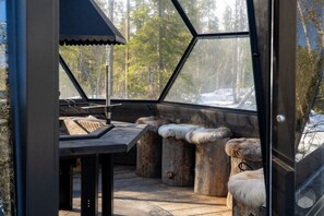 A wooden bench with fur seat cushions, a grill, and a view of a snowy forest through a glass enclosure. - North Suite Sky by Holiday in Lapland (Levi)
