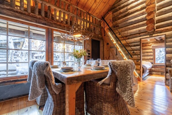 A log cabin interior with a dining table set for two, wicker chairs, a hanging light fixture, and a view of the snowy outdoors through large windows.