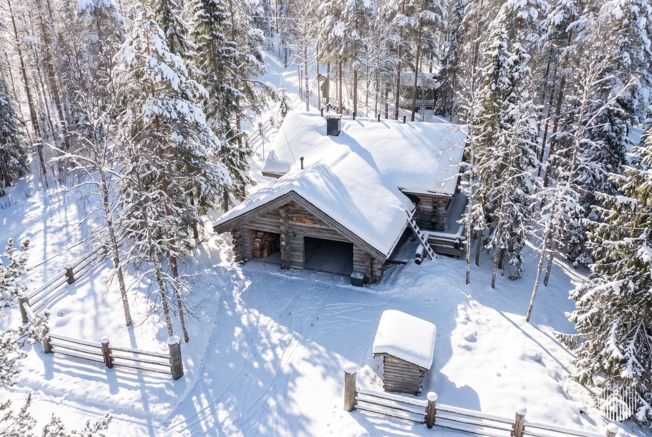 Una cabana de fusta amb un teulat cobert de neu, envoltada d'arbres i una tanca de fusta.