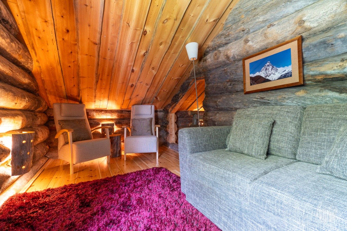 A log cabin interior with a wooden ceiling, a gray sofa, a red rug, and a framed picture on the wall.