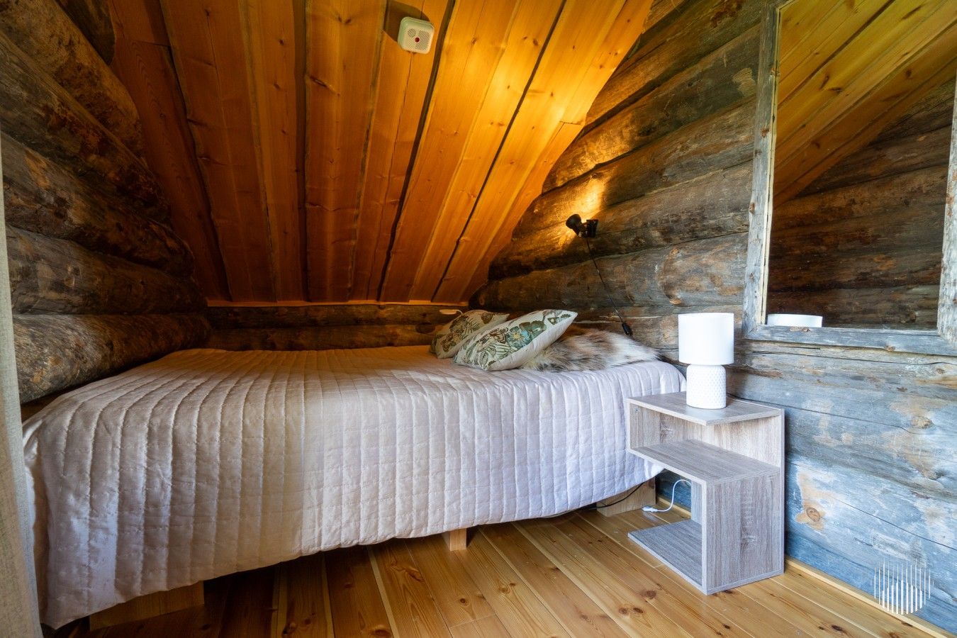 A single bed with a quilted bedspread and a pillow, a bedside table with a lamp, and a wooden floor in a log cabin room.