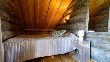 A single bed with a quilted bedspread and a pillow, a bedside table with a lamp, and a wooden floor in a log cabin room.