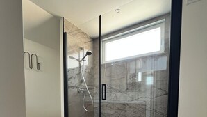 Un baño moderno con ducha envuelta en cristal, paredes de mármol y un pequeño cesto de basura.