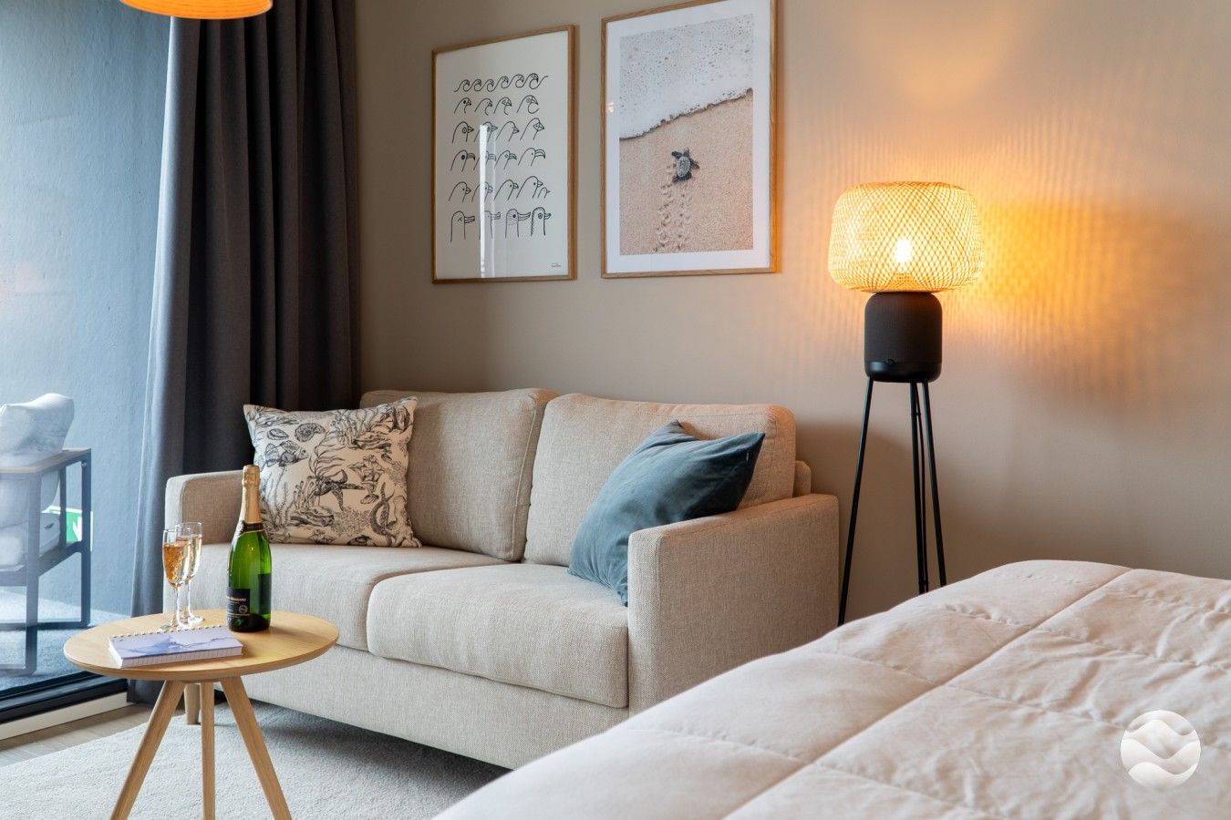 Hotellihuoneessa on sohva, yöpöytä, jossa on pullo ja lasi, lamppu sekä kaksi kehystettyä kuvaa seinällä.